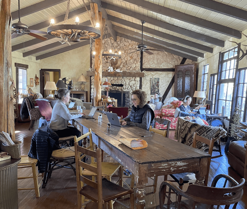 A bunch of writers sit working at table and in chairs at a lodge during a writing retreat.